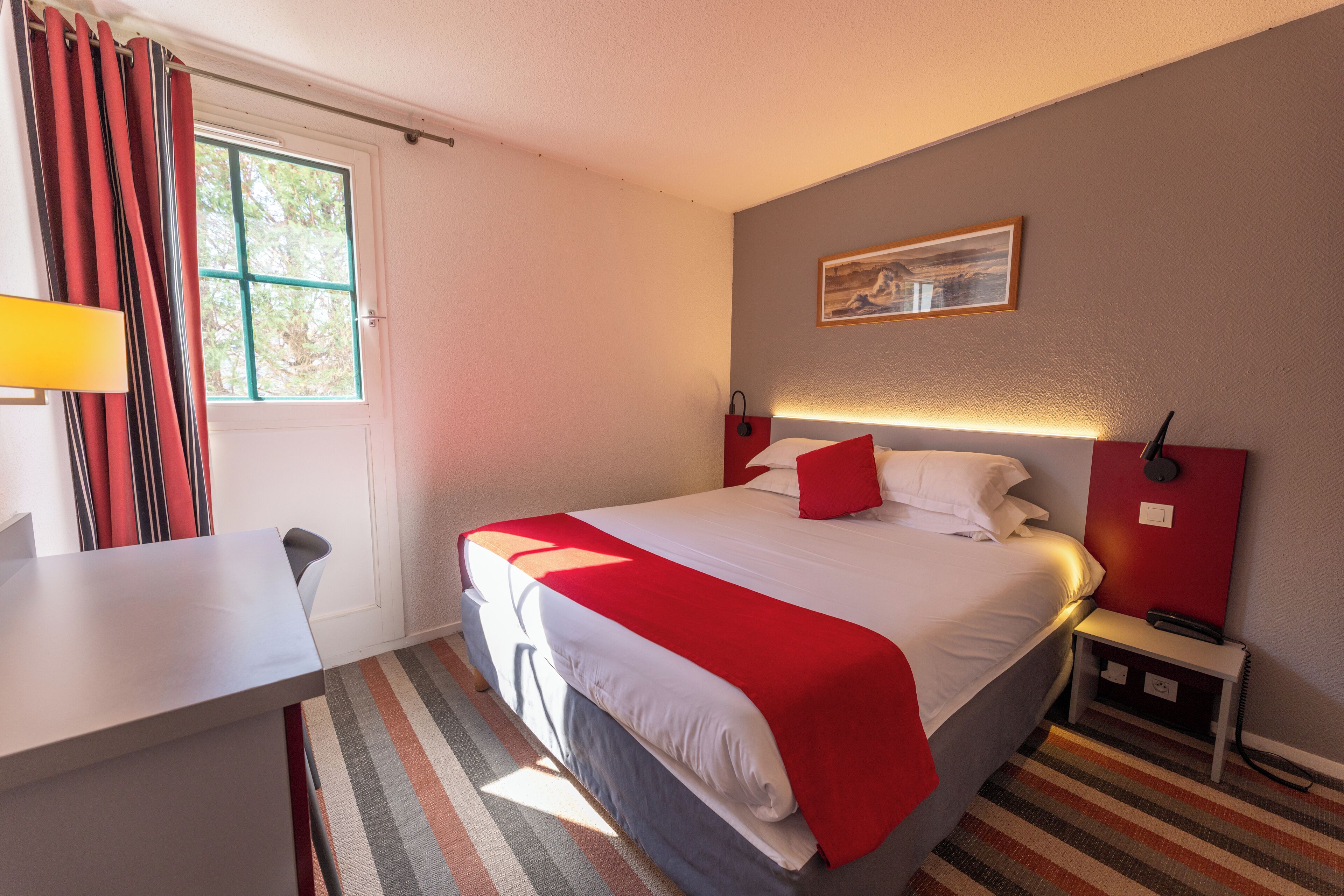 A hotel room with a bed, a desk, a chair, a nightstand, a lamp, and a window with curtains.