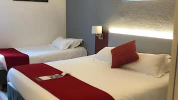 A hotel room with two beds, each with a red blanket and white linens. There is a framed picture on the wall above the beds and a lamp on the nightstand.