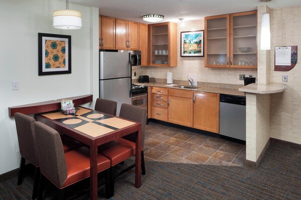 A compact kitchen with wooden cabinets, a refrigerator, microwave, stove, and dishwasher. There is a dining table with chairs and a patterned wall hanging.