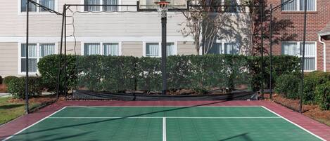 An outdoor basketball court with a net, surrounded by a fence and bushes, in front of a building with windows and a brick facade.
