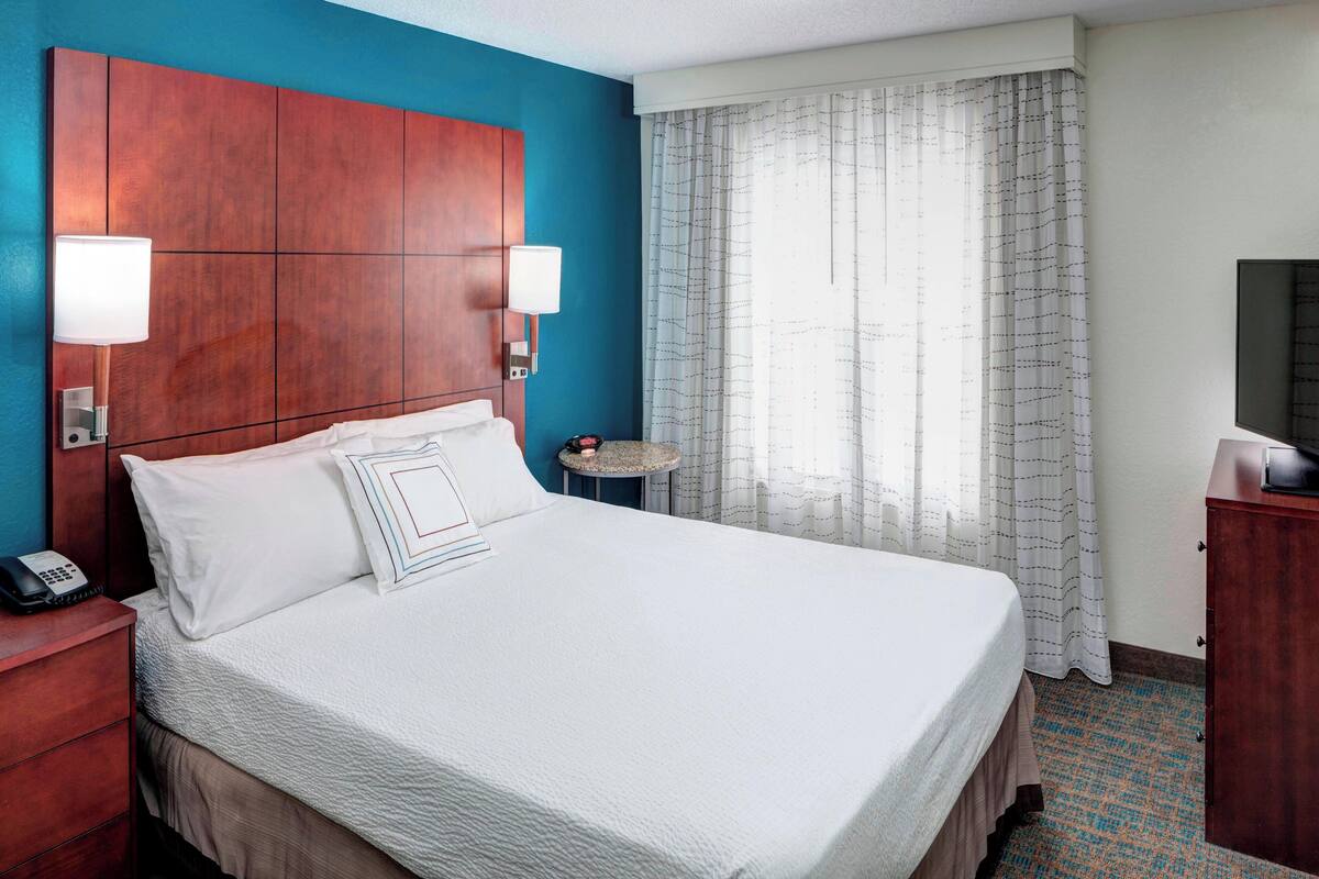 A hotel room with a large bed, a wooden headboard, two bedside lamps, a small round table, a telephone, and a television on a wooden cabinet.