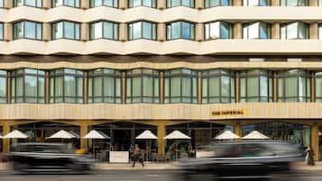 Ein Hotel mit einer modernen Fassade, zahlreichen Fenstern und einer Außenbestuhlung mit Sonnenschirmen.