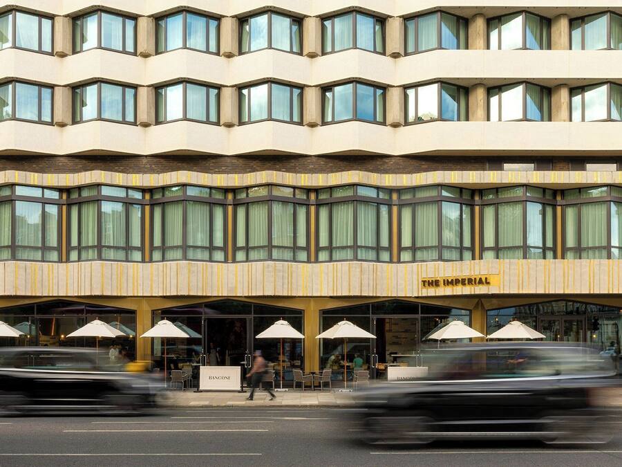 Ein Hotel mit einer modernen Fassade, zahlreichen Fenstern und einer Außenbestuhlung mit Sonnenschirmen.