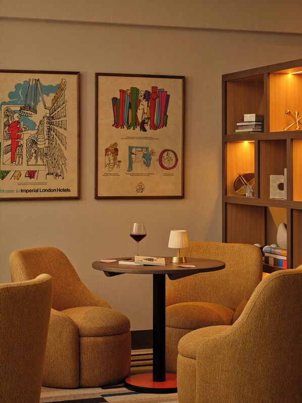 A hotel room with two armchairs, a small table with a wine glass and a lamp, and two framed artworks on the wall. - The Imperial (London)