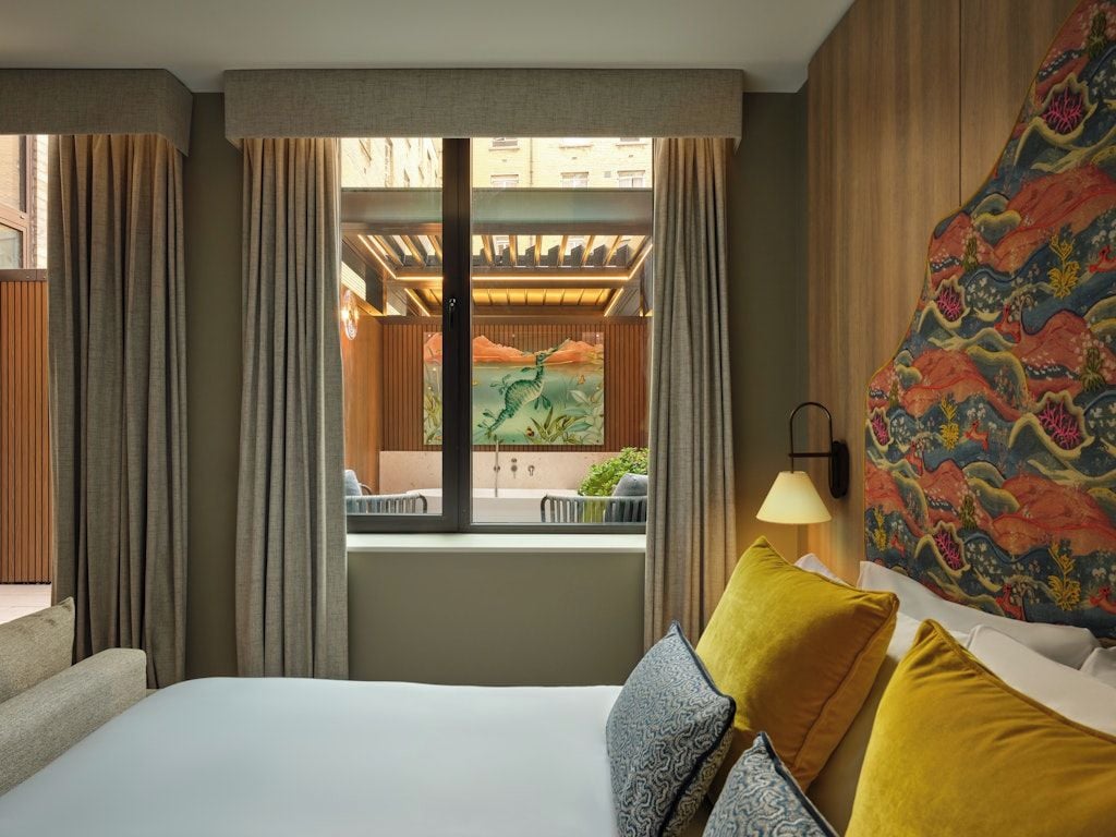 Una camera d'albergo con un letto, tende, vista su uno spazio esterno e un muro con un motivo colorato.