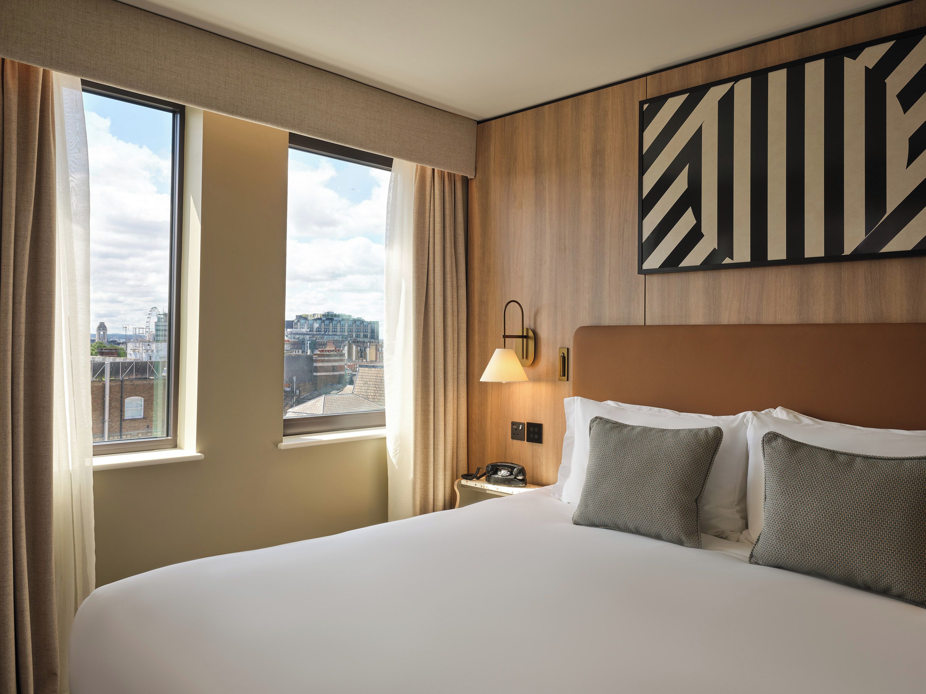 A hotel room with a large bed, a nightstand with a lamp, a view of the city through a window, and a wall-mounted artwork.