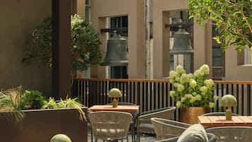 Uma área de estar ao ar livre com cadeiras e mesas de vime, cercada por plantas em vasos e vista para um edifício com grandes janelas.