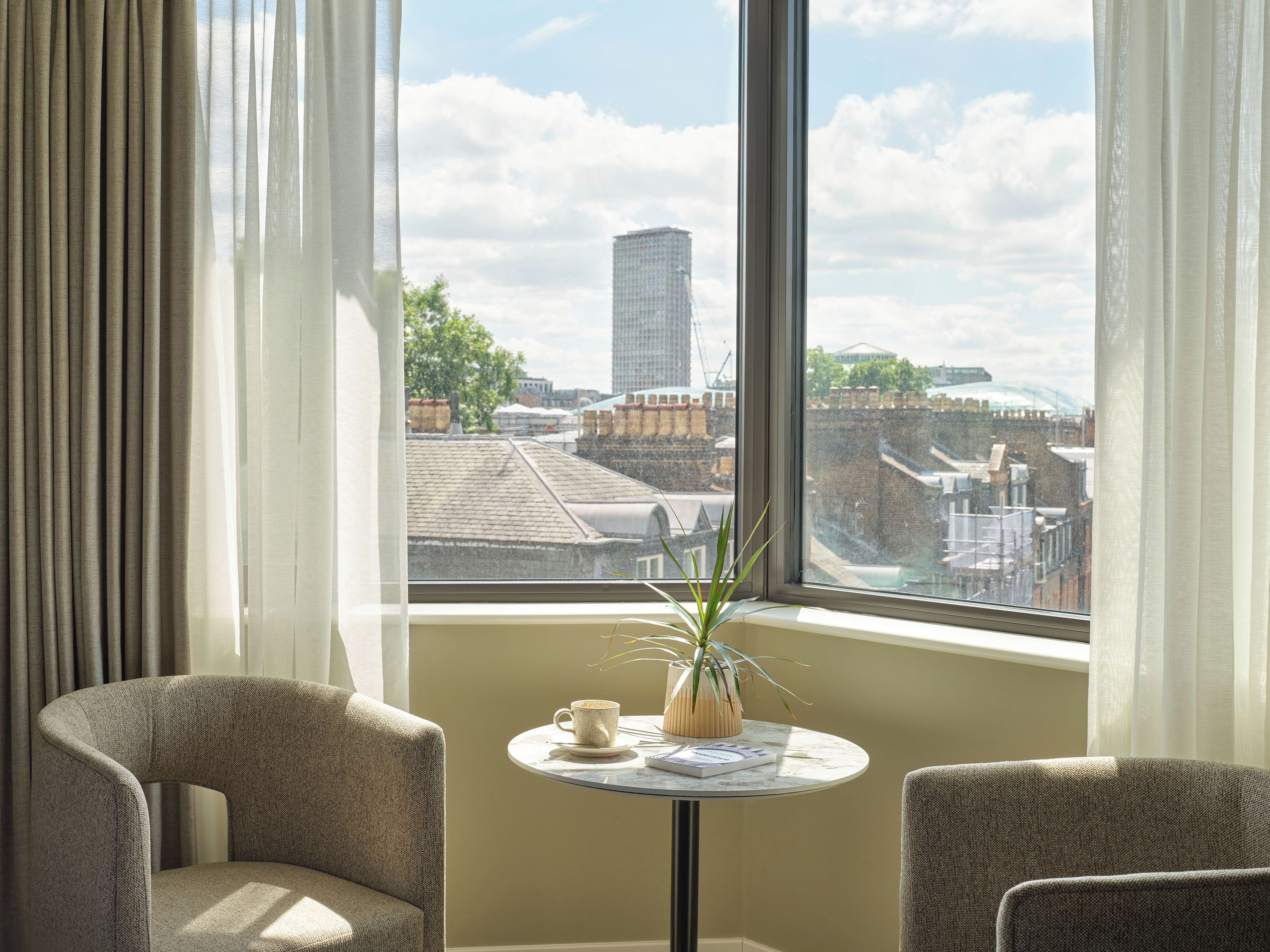 A room with a view of a cityscape, featuring a round table with a cup and a plant, and two chairs.