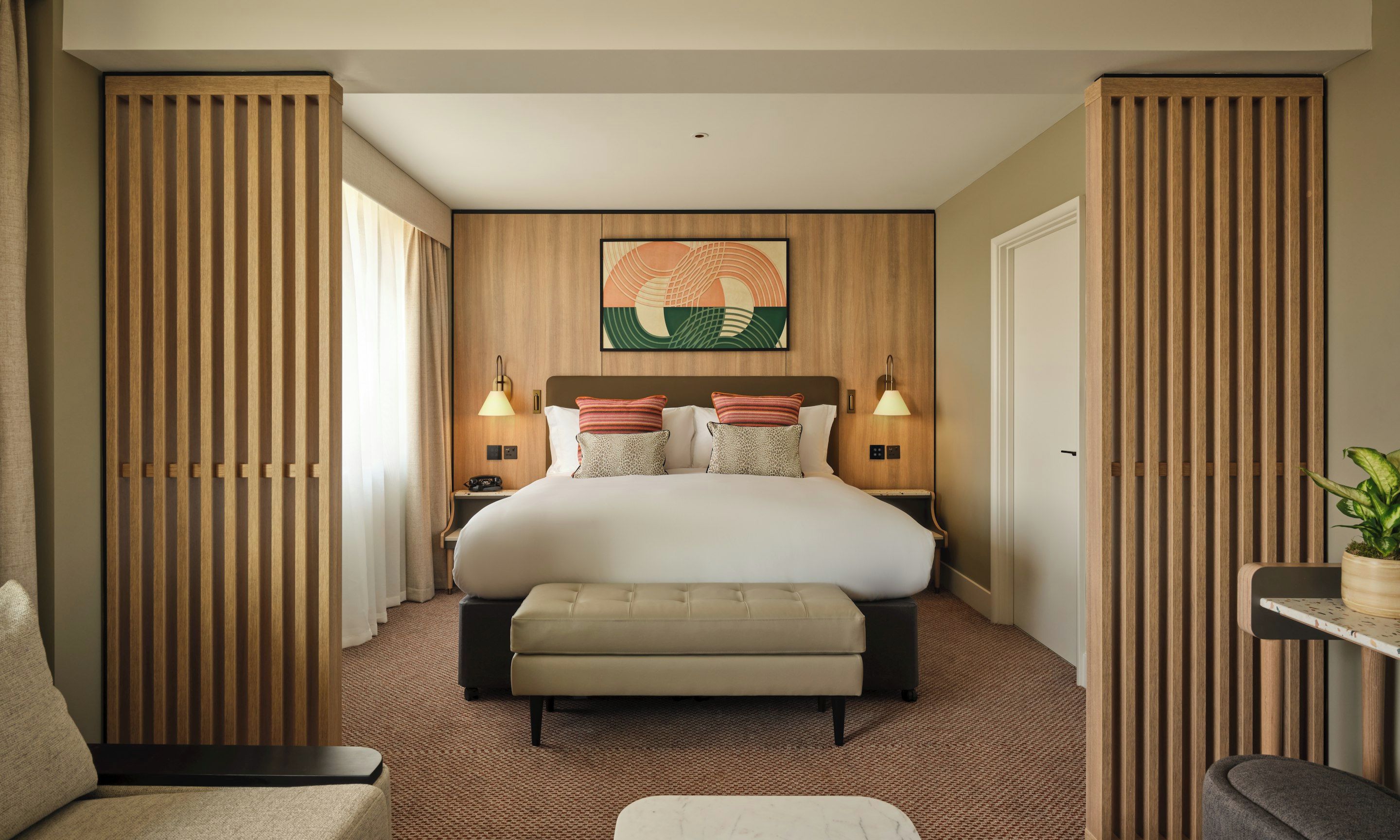A hotel room with a large bed, two bedside tables with lamps, a headboard with a decorative panel, a small bench, and a potted plant.