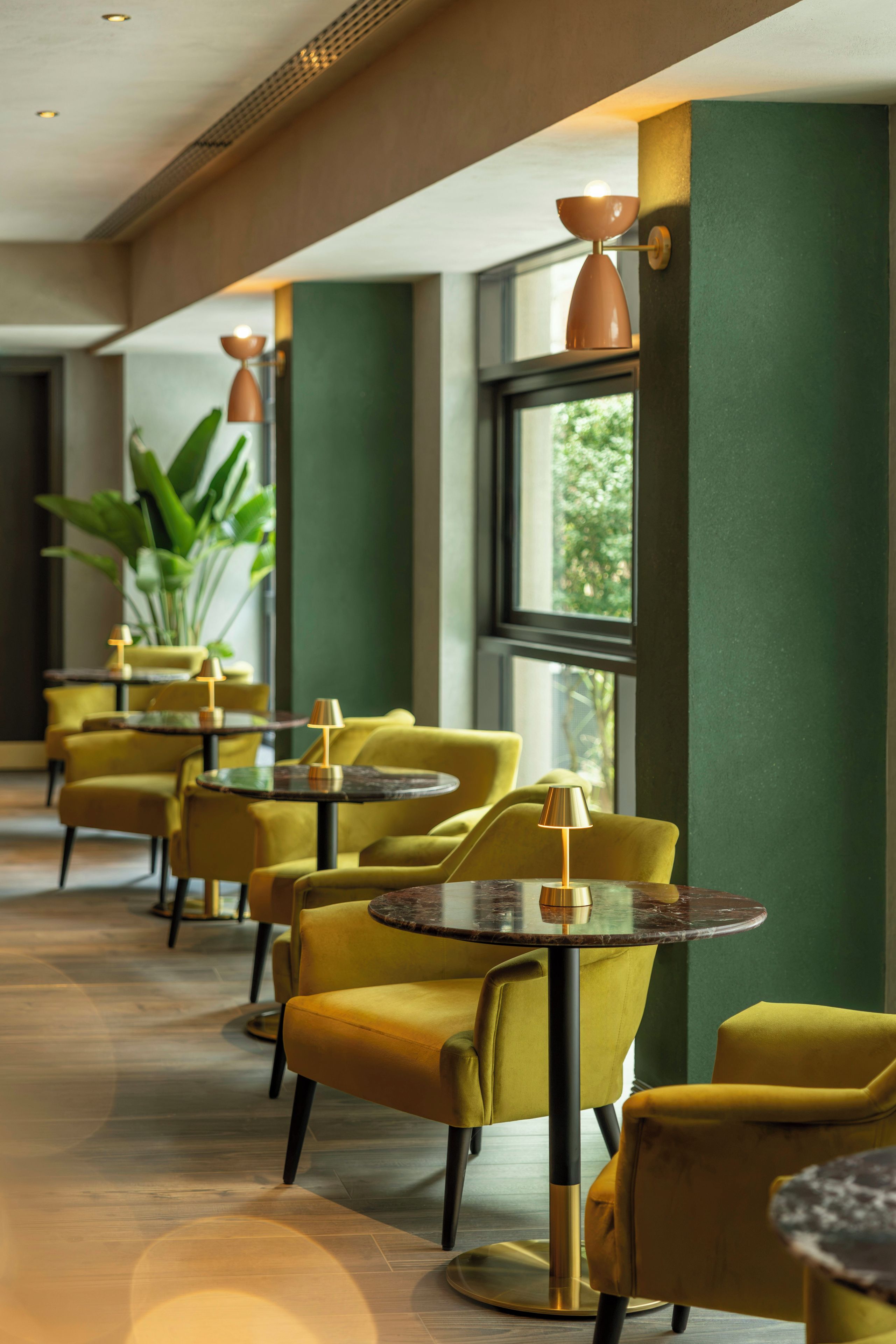 Et hotel lobby med gule stole og runde borde, store vinduer og grønne vægge.