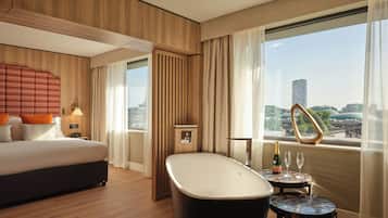 Une chambre d’hôtel moderne avec un grand lit, une baignoire, une petite table avec une bouteille et un verre, et une vue sur le paysage urbain.
