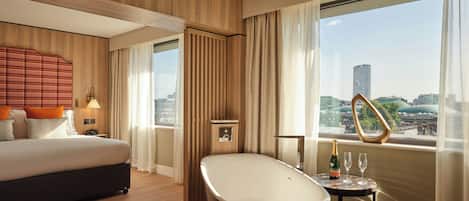 A modern hotel room with a large bed, a bathtub, a small table with a bottle and glass, and a view of the cityscape.