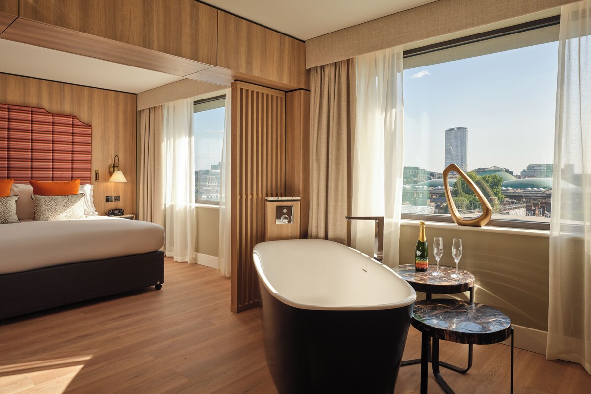Ein modernes Hotelzimmer mit einem großen Bett, einer Badewanne, einem kleinen Tisch mit einer Flasche und einem Glas sowie Blick auf die Stadt.