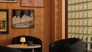 En hotellobby med ett runt bord, två fåtöljer, en lampa, inramade tavlor och en vägg med en prislista.