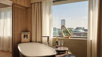 A modern bathroom with a freestanding bathtub, a small round table with a bottle and glasses, and a view of a cityscape through a large window.