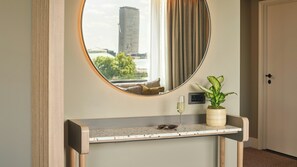 Una camera d'albergo con uno specchio rotondo, un tavolino con una pianta in vaso, un bicchiere da vino e vista su un edificio attraverso la finestra.
