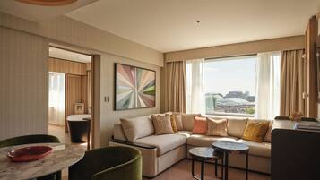 A modern hotel room with a sectional sofa, a dining area with a round table, and a large window offering a city view.