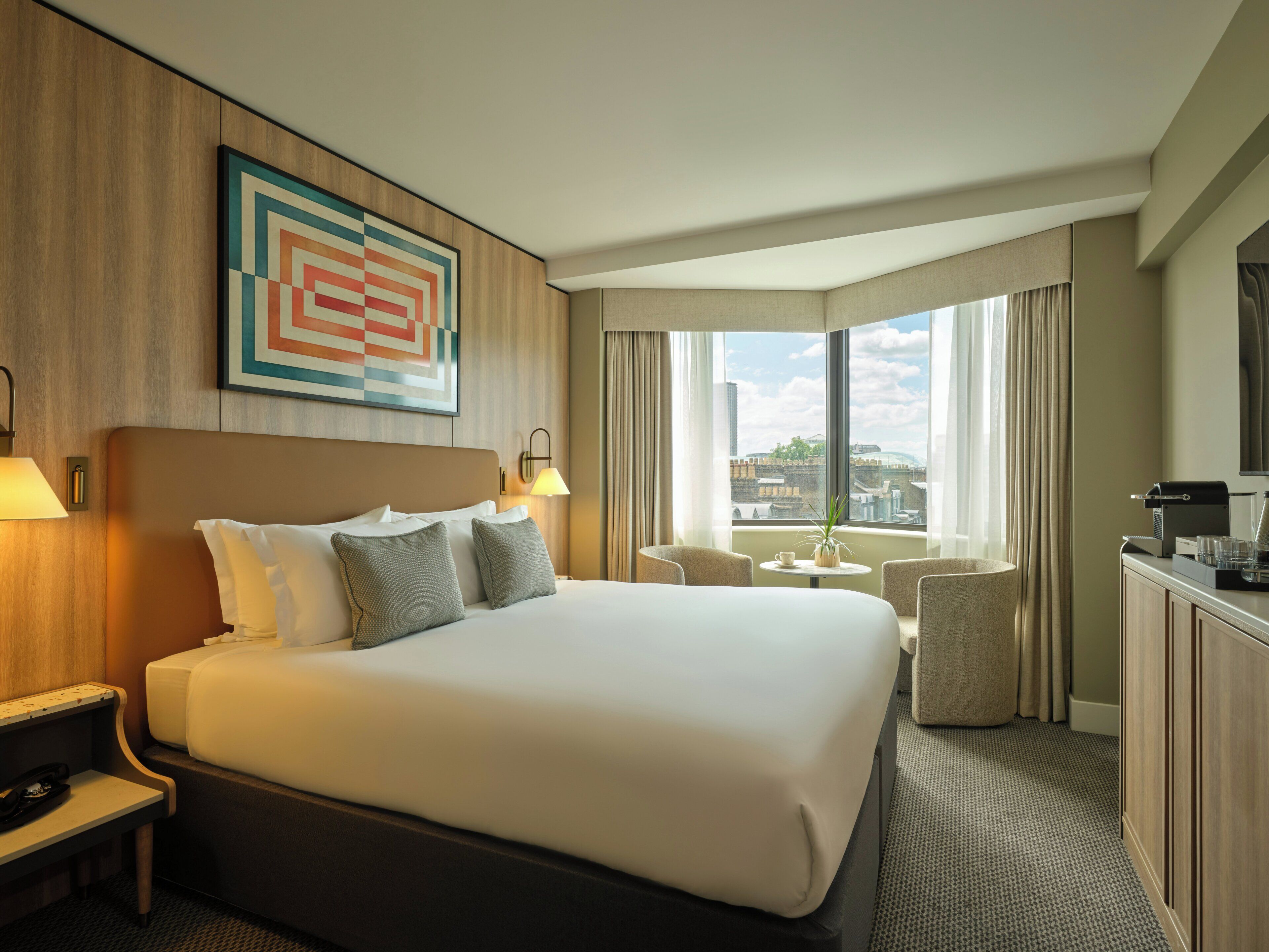 A hotel room with a large bed, a desk, a chair, a small table with a plant, and a view of the city through the window.
