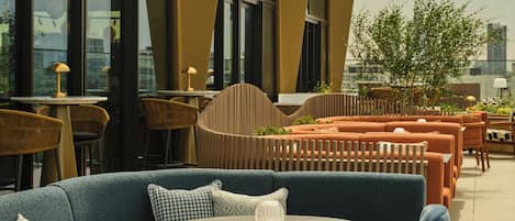 An outdoor seating area with round tables and blue cushioned chairs, large windows, and a modern architectural design.
