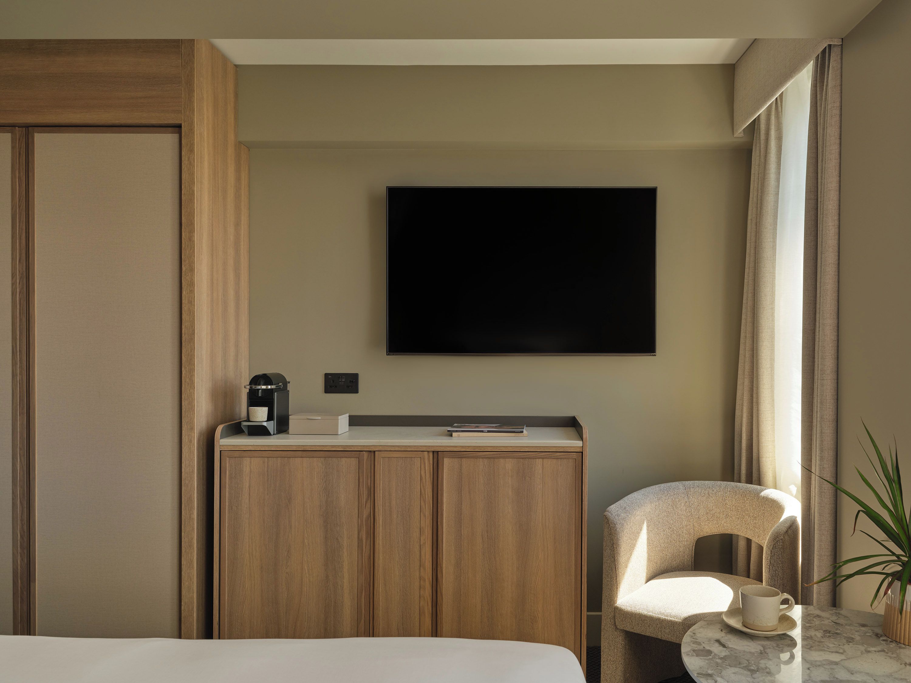 A hotel room with a bed, a desk with a coffee maker, a TV mounted on the wall, and a chair with a cup on a table.