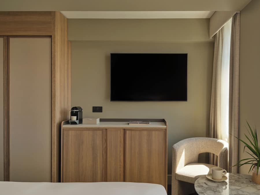 A hotel room with a bed, a desk with a coffee maker, a TV mounted on the wall, and a chair with a cup on a table.