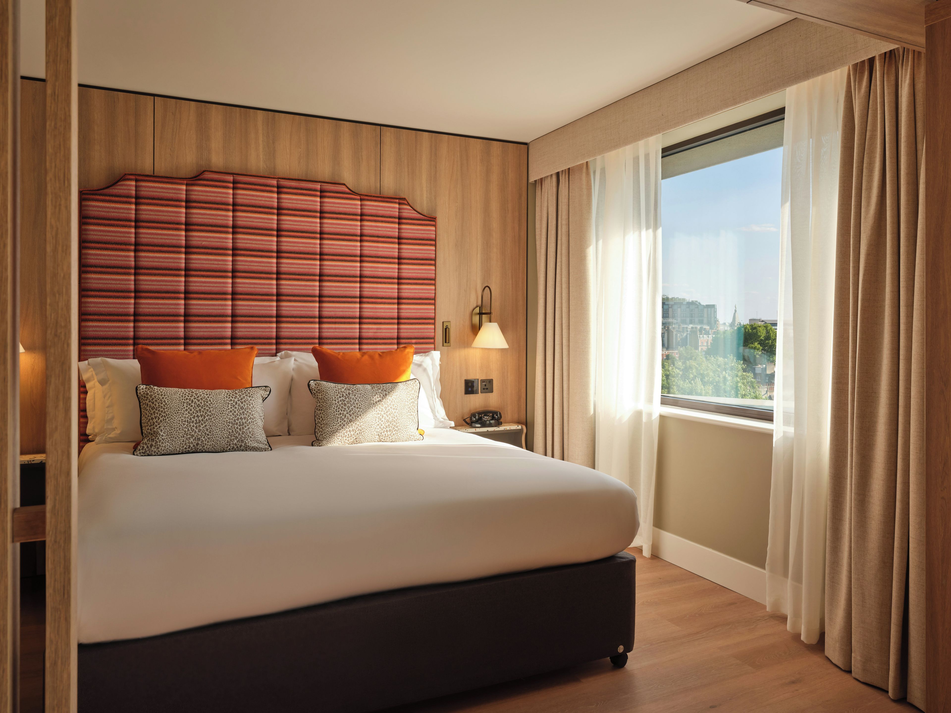 Quarto de hotel com uma cama grande, cabeceira, mesa de cabeceira, abajur e uma janela com cortinas.