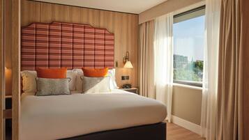 Quarto de hotel com uma cama grande, cabeceira, mesa de cabeceira, abajur e uma janela com cortinas.
