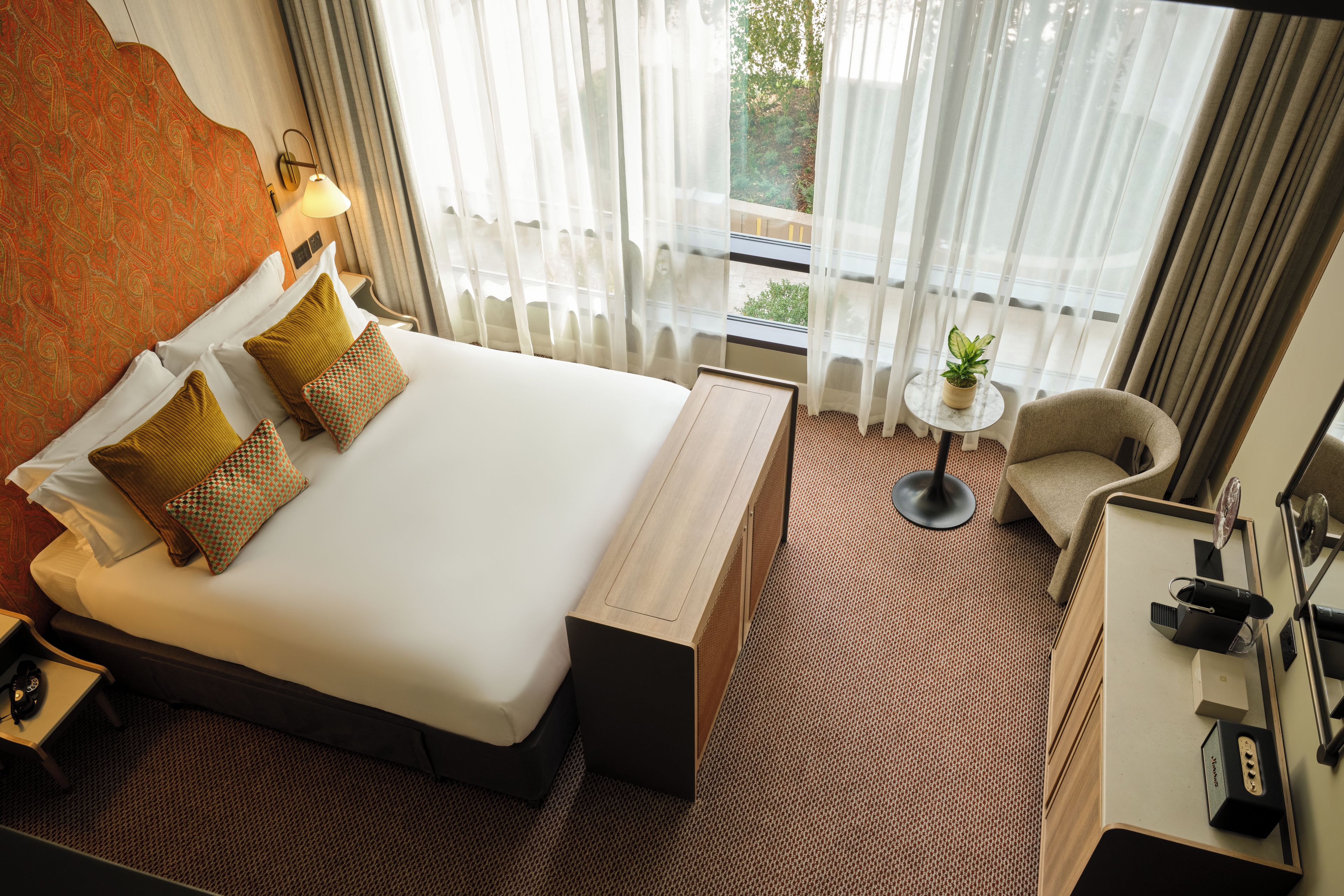 Quarto de hotel com uma cama grande, mesa de cabeceira, uma cadeira, pequena mesa com planta e vista para o exterior através de cortinas finas.