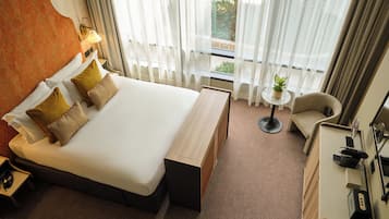Quarto de hotel com uma cama grande, mesa de cabeceira, uma cadeira, pequena mesa com planta e vista para o exterior através de cortinas finas.