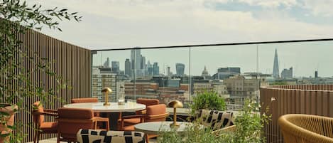 Una terraza en la azotea con vistas al horizonte urbano, amueblada con mesas redondas, sillas y plantas en macetas.