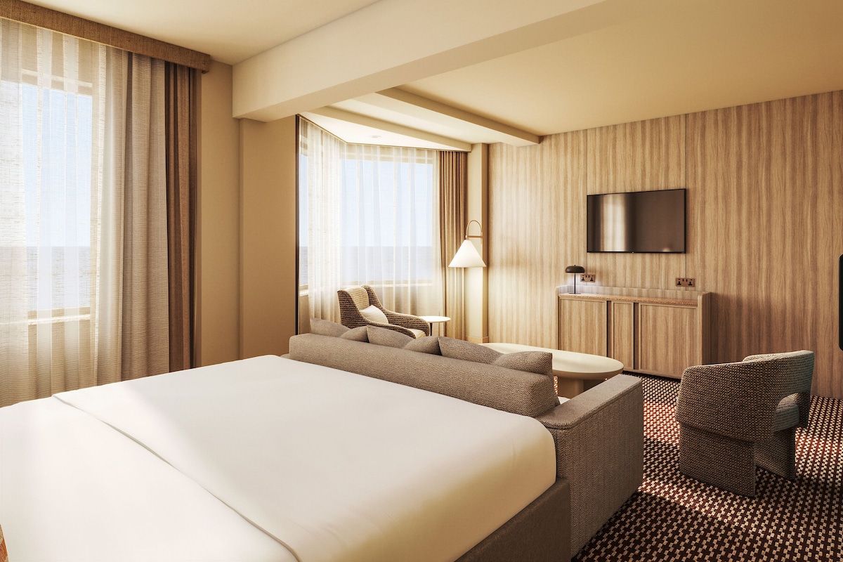 A hotel room with a large bed, a seating area with a chair and table, a TV mounted on the wall, and a window with curtains.