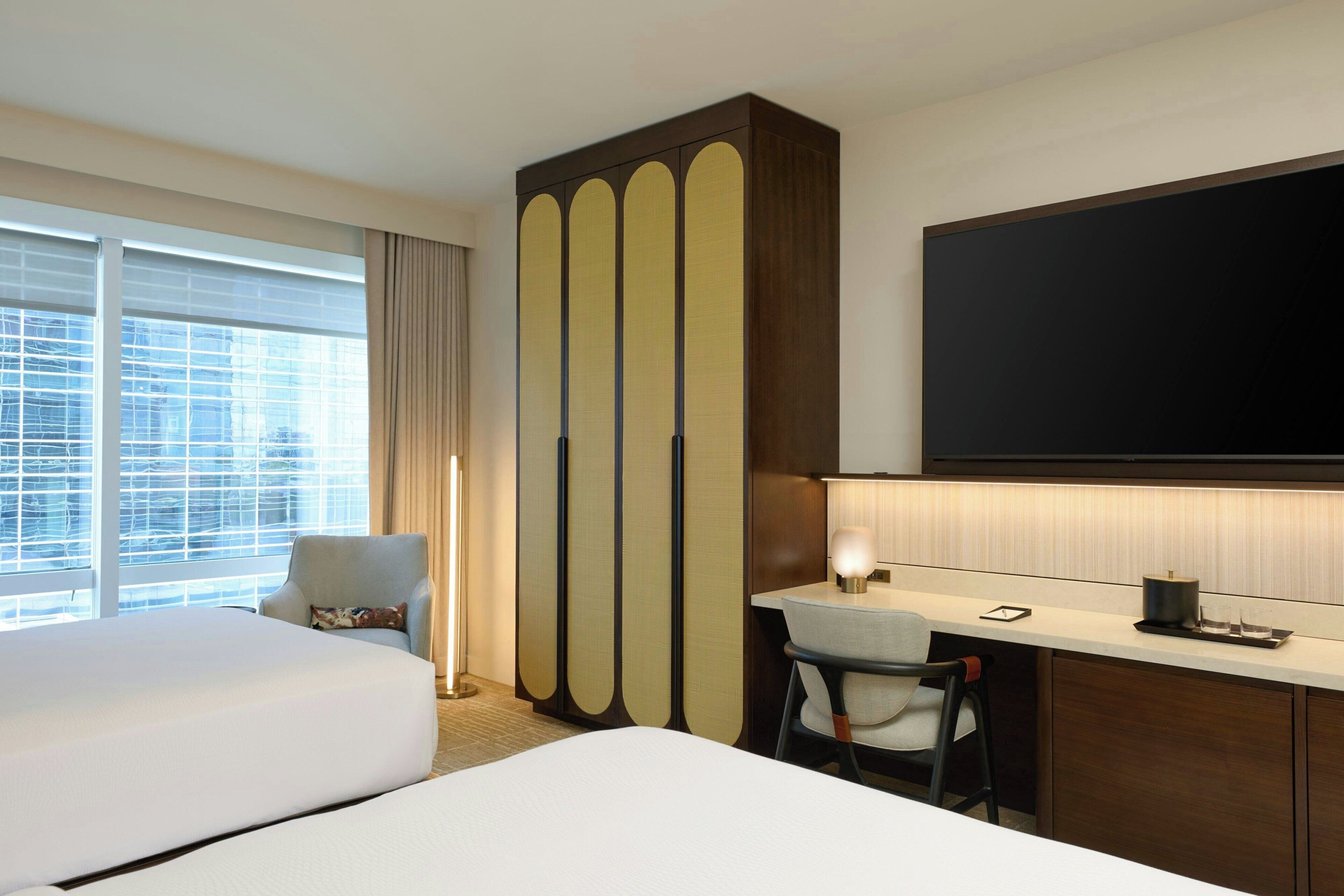 Habitación de hotel con dos camas, un escritorio, una silla, un televisor y un ventanal con cortinas.