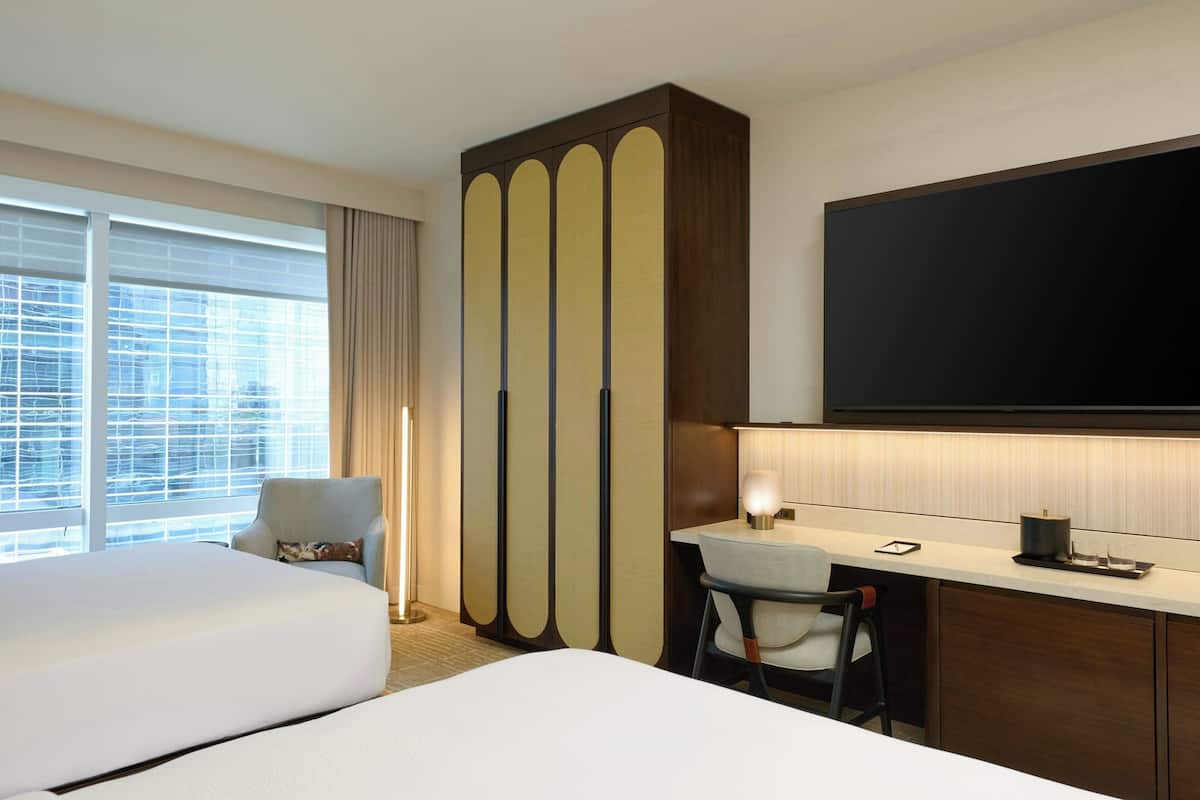 Habitación de hotel con dos camas, un escritorio, una silla, un televisor y un ventanal con cortinas.