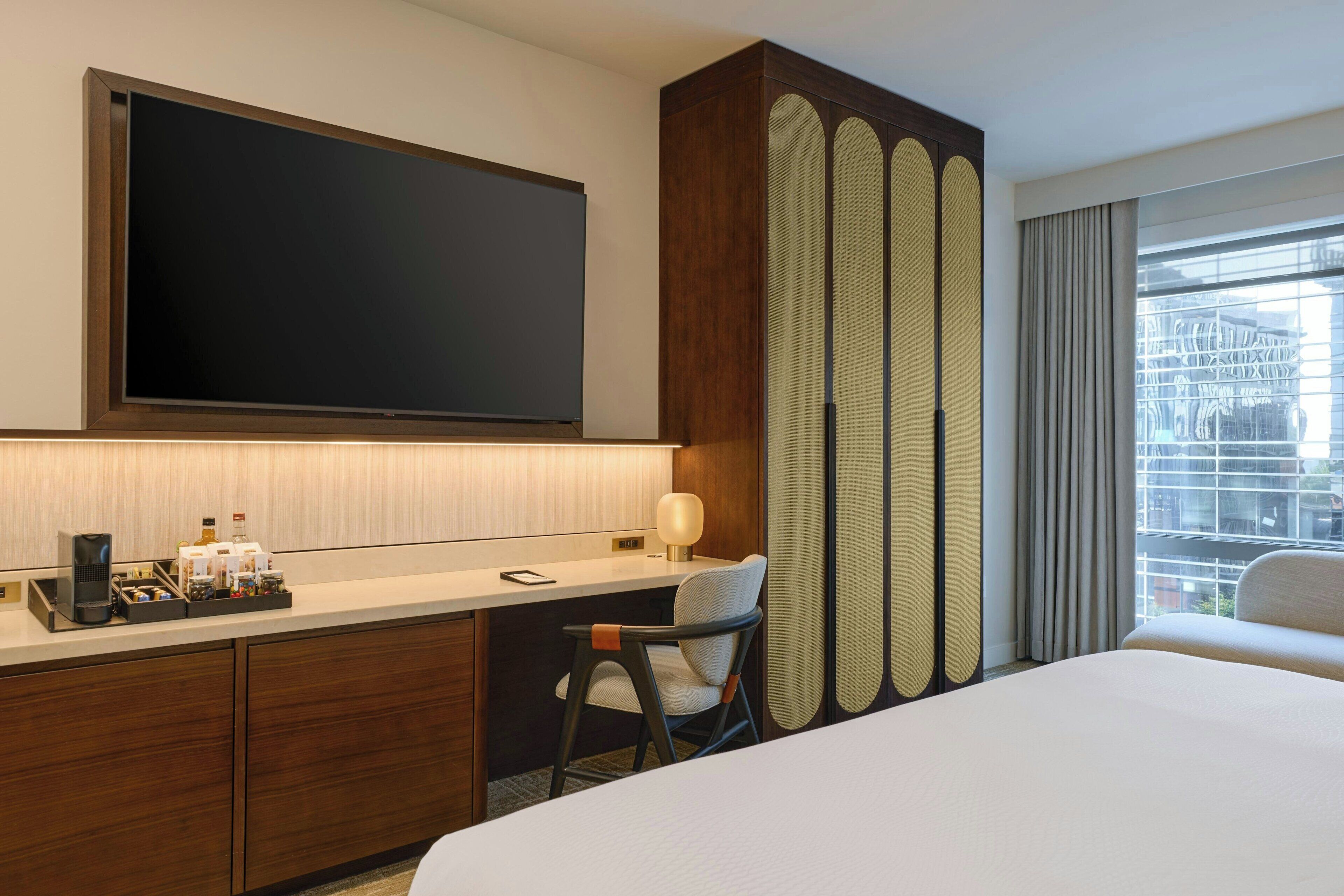 A hotel room with a bed, a desk with a computer, a chair, a TV, and a window with curtains.
