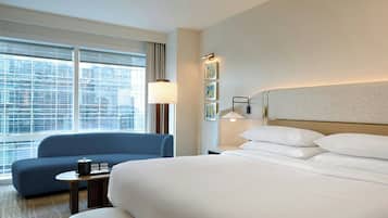A hotel room with a large bed, a blue armchair, a small table with a lamp, and a view of the city through the window.