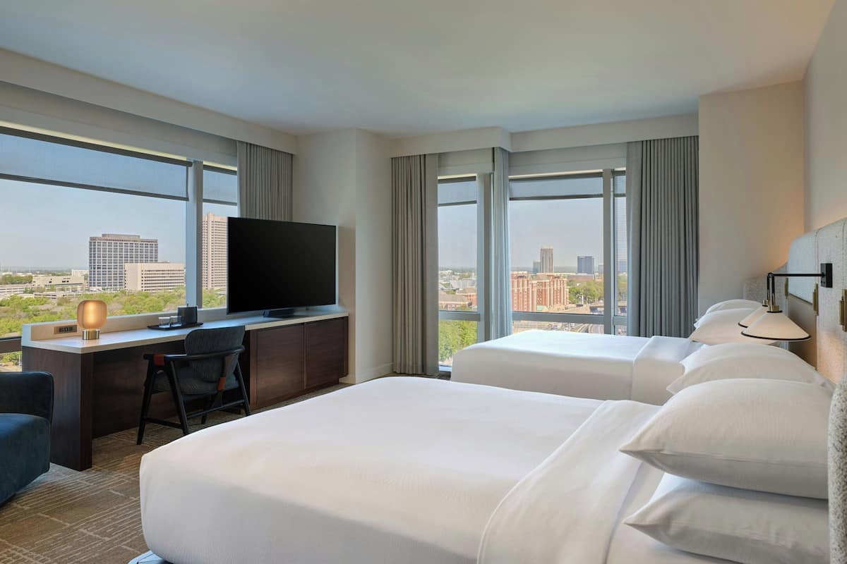 Habitación de hotel con escritorio, televisor, dos camas y ventanas amplias con vistas a la ciudad.