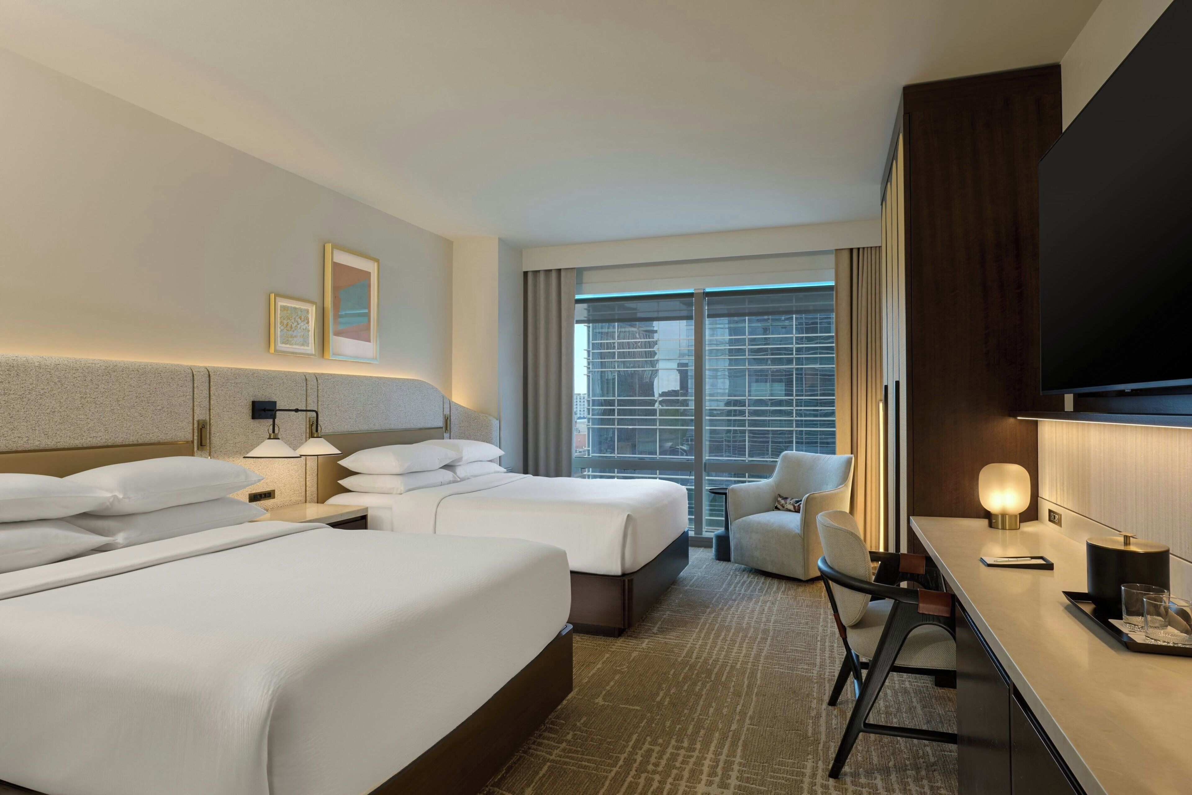 A hotel room with two beds, a desk, a chair, a TV, and a large window with a view of buildings.