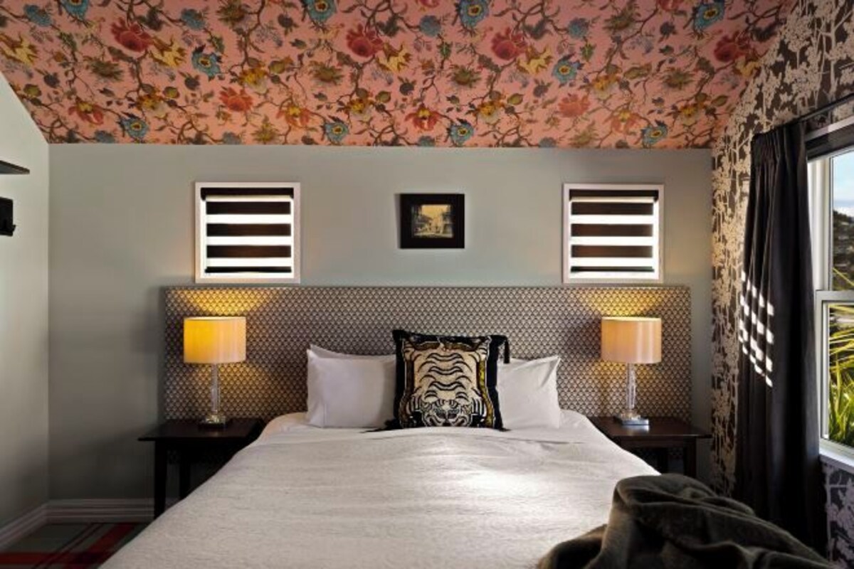 A hotel room with a bed, two bedside lamps, a headboard with a patterned design, a window with curtains, and a floral ceiling.
