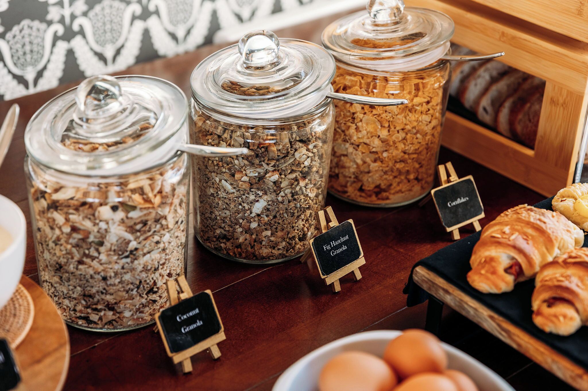 A breakfast buffet with granola jars, a bowl of eggs, and croissants.