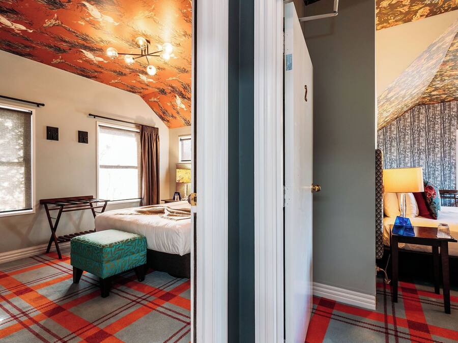 A room with a plaid carpet, a bed with a white duvet, a bench, and a table with a lamp.