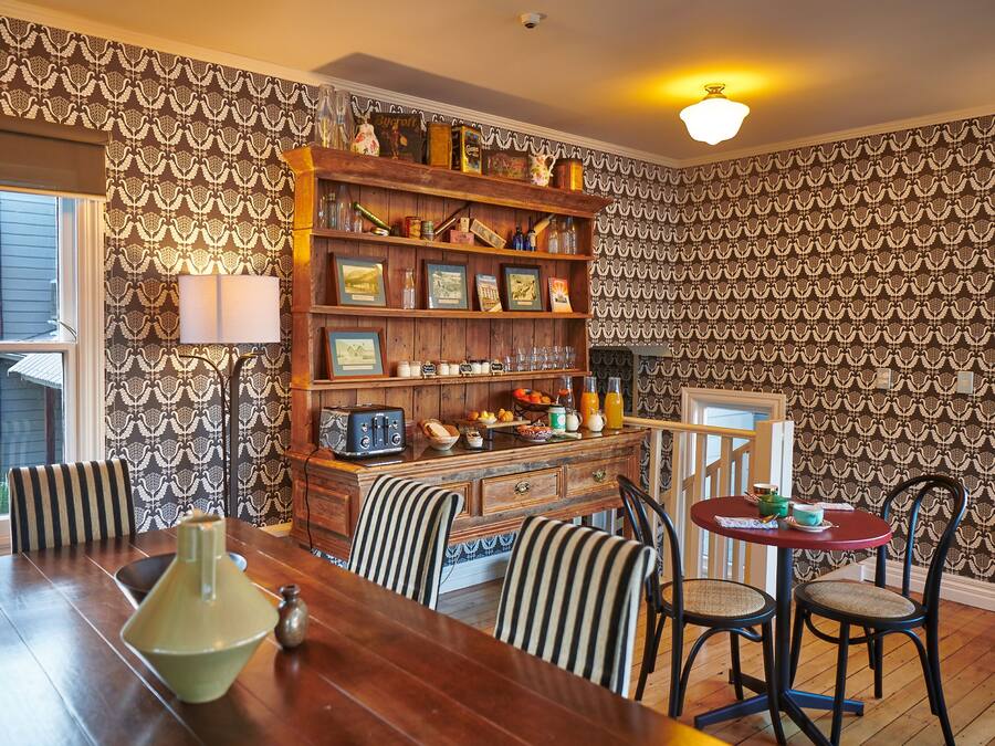 Un comedor con una mesa de madera, sillas a rayas, una estantería y un papel tapiz estampado.