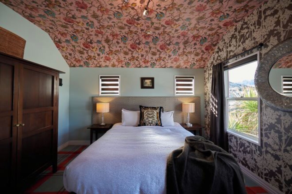 A bedroom with a floral ceiling, a bed with white bedding, two bedside lamps, a nightstand, a wooden wardrobe, and a window with a view.