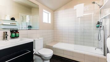A modern bathroom with a white sink, toilet, and bathtub. There is a window providing natural light, and toiletries are visible on the counter.