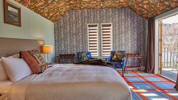 A bedroom with a large bed, a desk, two chairs, a window with shutters, and a balcony with a railing.