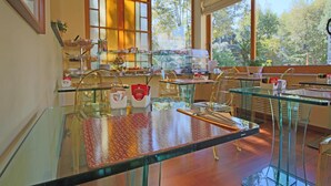 A breakfast buffet with glass tables and chairs, a variety of food items, and a view of trees through large windows. - Il Giardino di Albaro (Genoa)