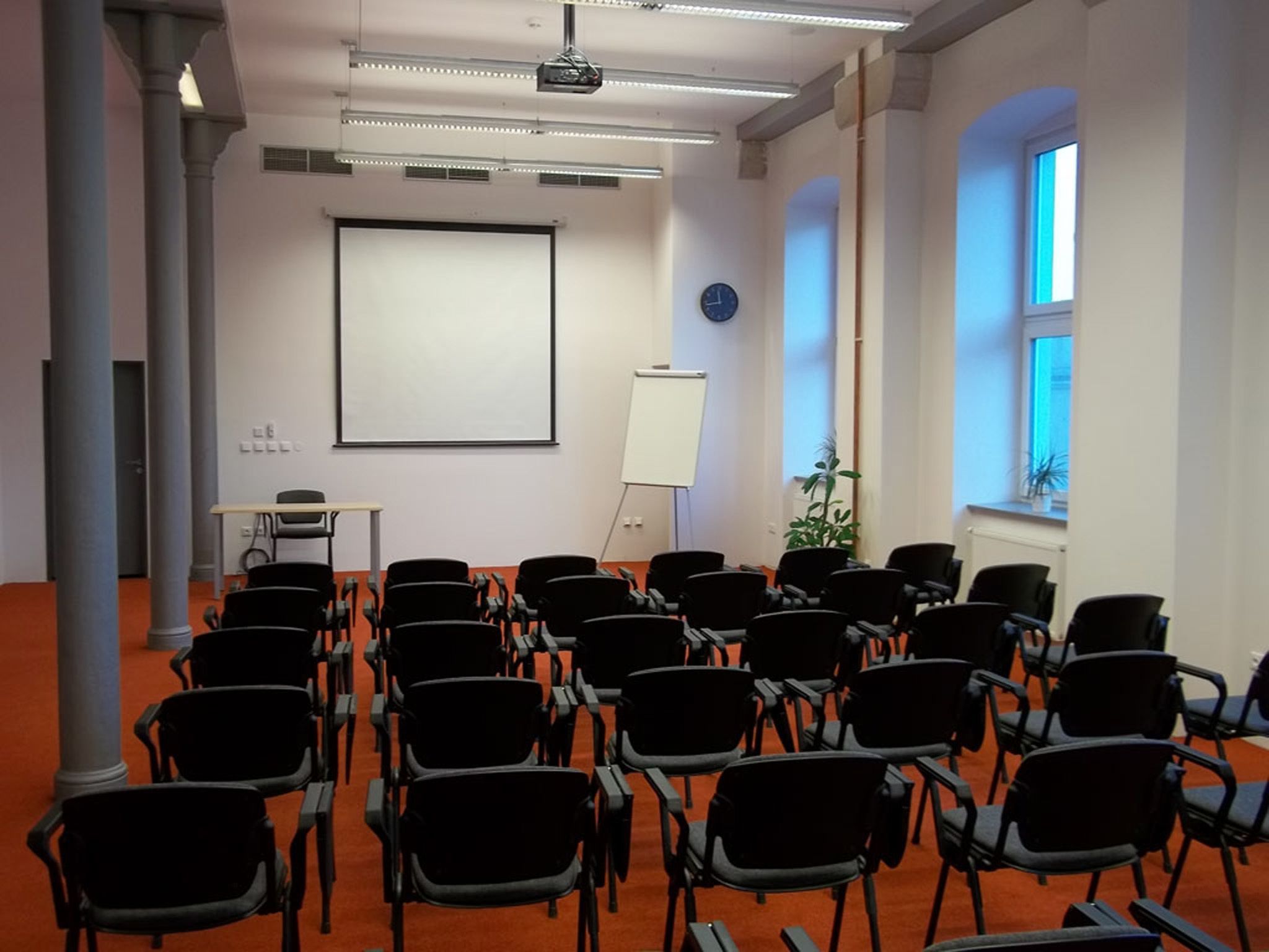 A conference room with rows of black chairs, a whiteboard, a projector screen, and a clock on the wall.