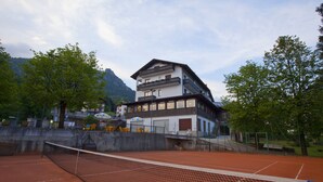 Una cancha de tenis con red, rodeada de árboles y un edificio con balcones.