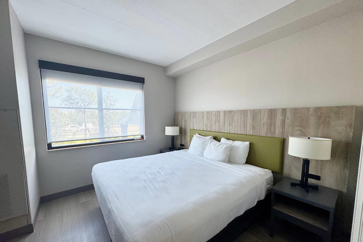 Quarto de hotel com uma cama grande, duas mesas de cabeceira com luminárias, uma janela com persianas e uma cabeceira de madeira.