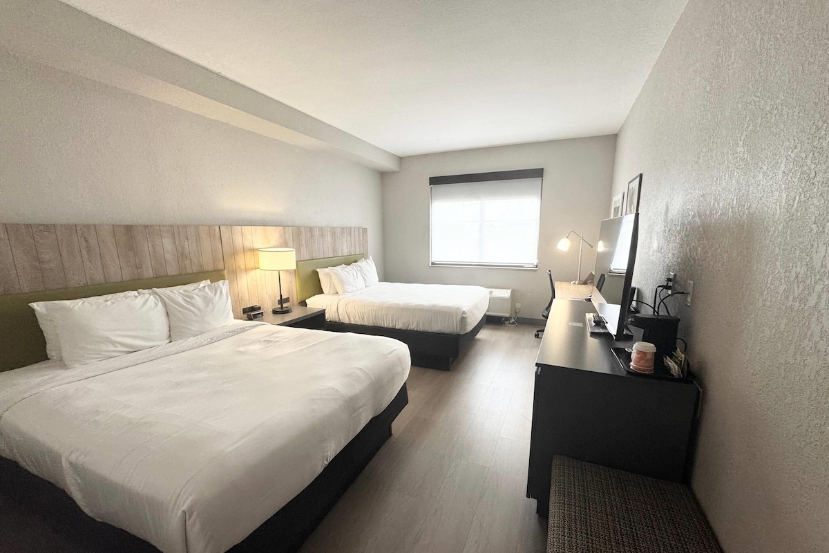 Quarto de hotel com duas camas, uma escrivaninha, uma cadeira, um abajur, uma janela e uma televisão de tela plana.