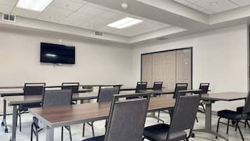 A conference room with a long table, multiple chairs, a flat-screen TV, ceiling lights, and a door.